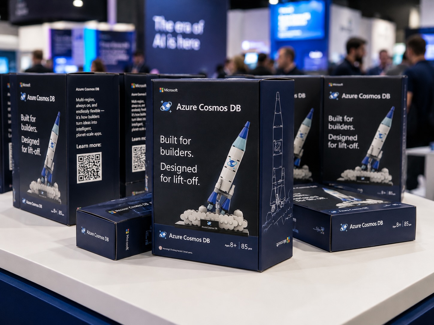 Navy Azure Cosmos DB Lego set boxes on a white booth counter, each showing a white and blue rocket lifting off from brick clouds.