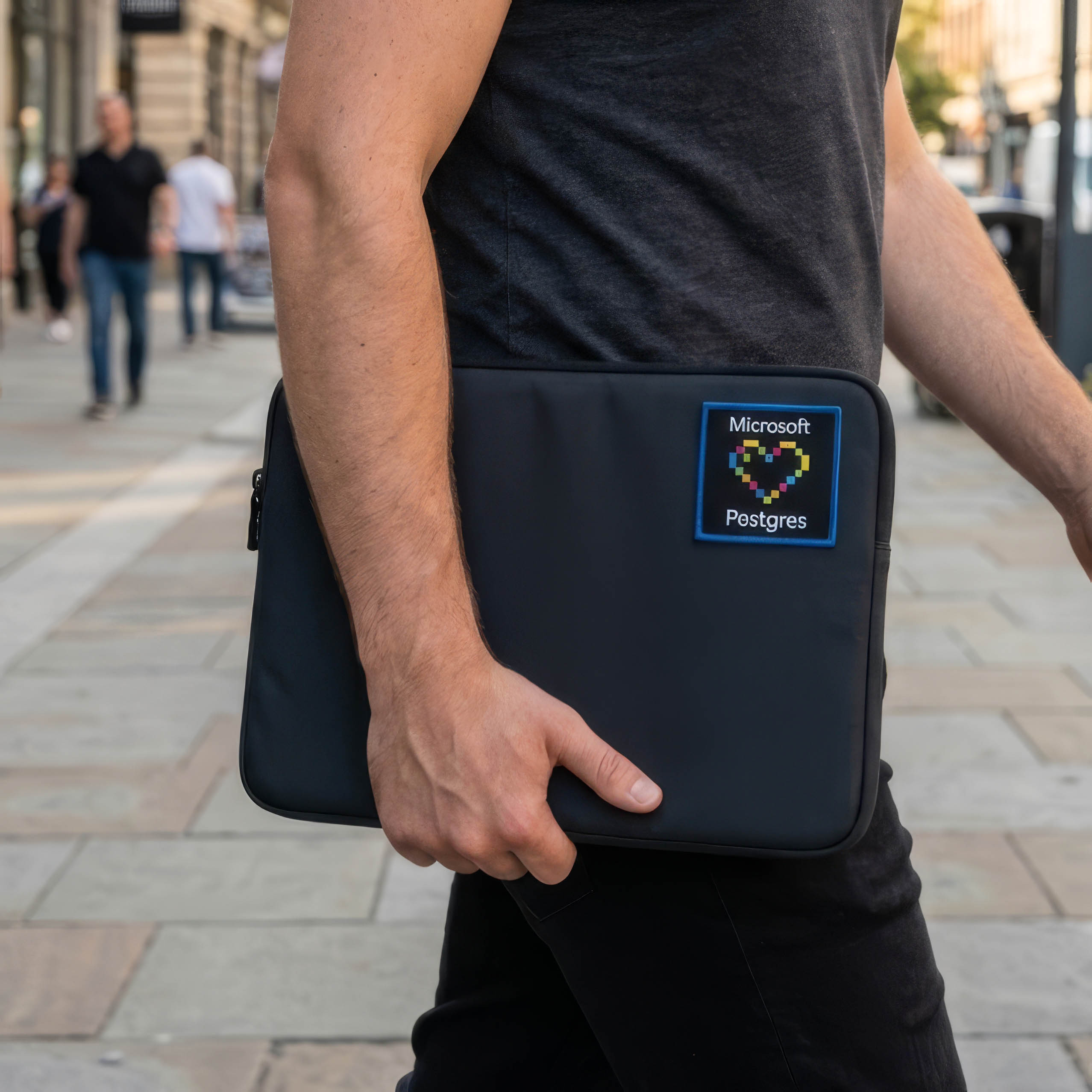 Person carrying a black laptop sleeve with a square Microsoft Postgres patch showing a pixel heart.
